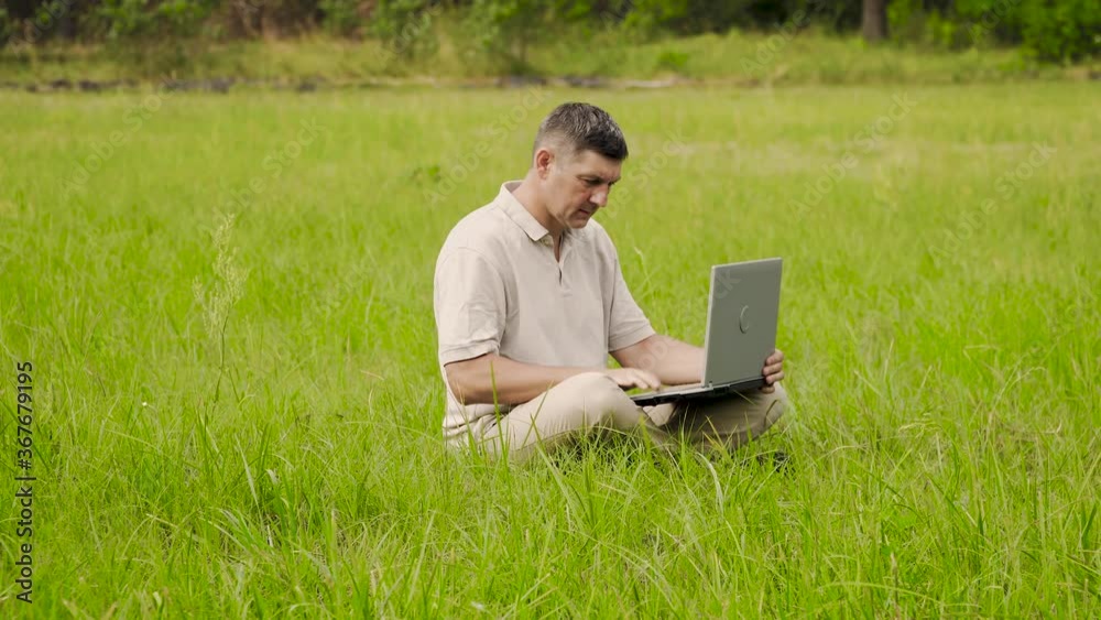 Man Works behind a Laptop Sitting on the Grass At The Sunny Day. Freelancer Remote Work Concept. 4K Man Works behind a Laptop Sitting on the Grass At The Sunny Day. Freelancer Remote Work Concept. 4K
