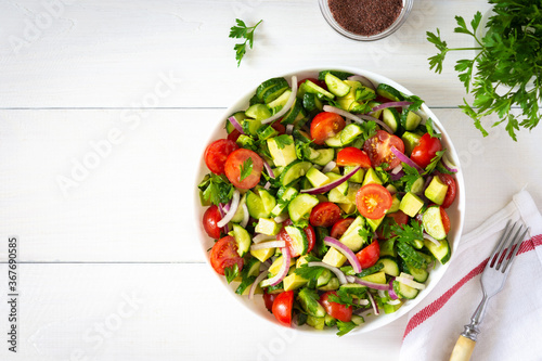 Salad of fresh ripe tomato, onion, avocado, cucumber and parsley and olive oil, salad in a white plate with a fork and towel on a white wooden background