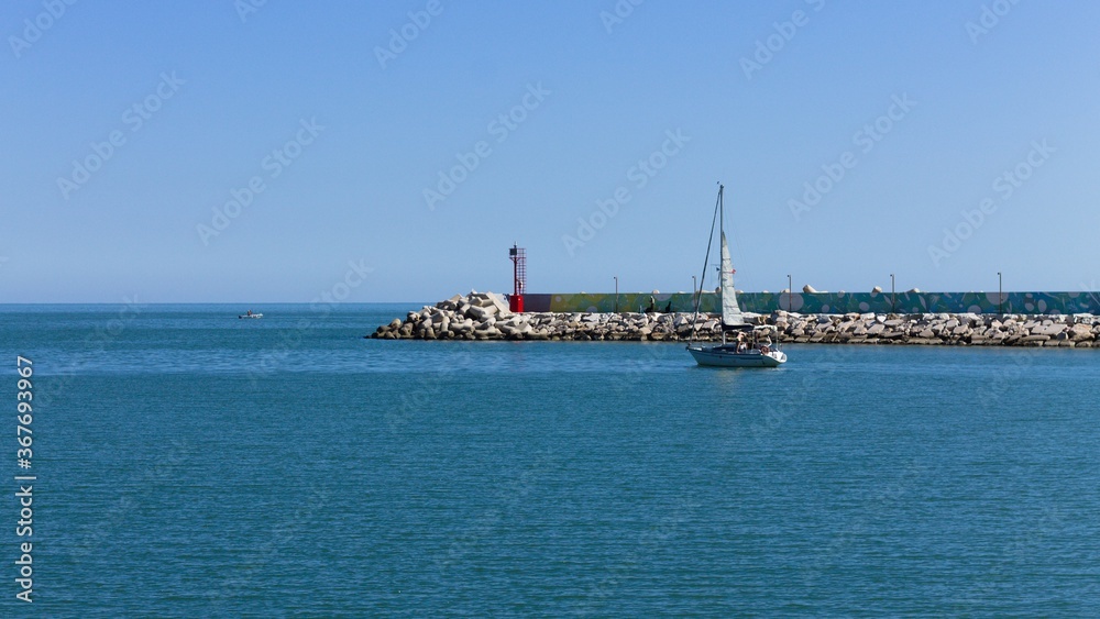 A little boat is sailing in front of the pier of Pesaro harbor with breakwaters and a small red lighthouse (Pesaro, Italy, Europe)