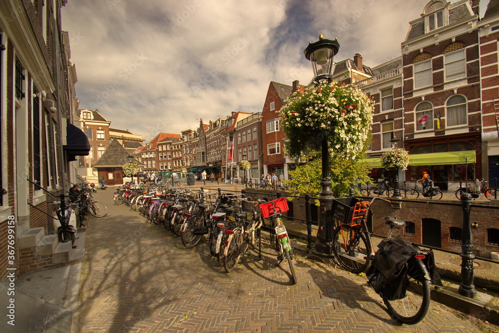 Ein Straße an einem Gracht in Utrecht mit viele Häuser, vielen Fahrräder und Strassen Laternen mit unzählige weiße und rote Blumen unter dem blauen Himmel mit weißen Wolken.