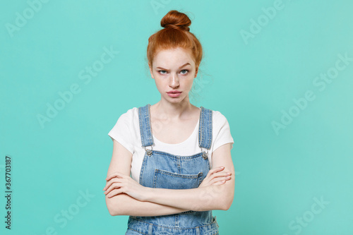 Perplexed displeased young readhead girl in casual denim clothes posing isolated on blue turquoise wall background studio portrait. People lifestyle concept. Mock up copy space. Holding hands crossed.