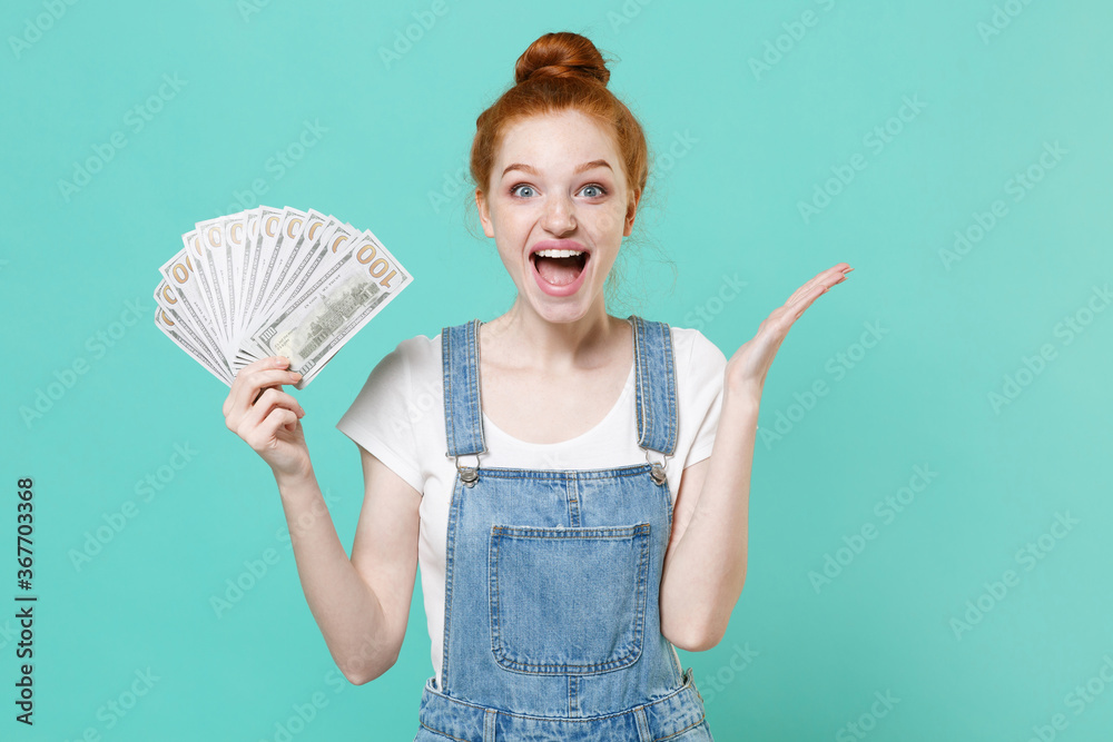 Excited young readhead girl in casual denim clothes isolated on blue turquoise background. People lifestyle concept. Mock up copy space. Hold fan of cash money in dollar banknotes, spreading hands. Excited young readhead girl in casual denim clothes isolated on blue turquoise background. People lifestyle concept. Mock up copy space. Hold fan of cash money in dollar banknotes, spreading hands.