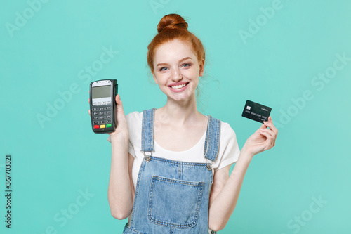 Funny young readhead girl in casual denim clothes isolated on blue turquoise background. People lifestyle concept. Hold wireless modern bank payment terminal to process acquire credit card payments.