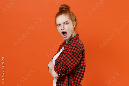 Side view of shocked irritated young readhead girl in casual red checkered shirt isolated on orange wall background. People lifestyle concept. Mock up copy space. Holding hands crossed looking camera.