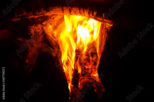 Big tree trunks, logs are burning into bonfire in dark on nature. Branches burns in fireplace in campsite. Bright orange tongues of flame among firewoods and red coals. Resting in campint outdoors.