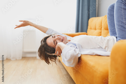 Joyful funny young woman girl in casual clothes lying on couch spending time in living room at home. Rest relax good mood leisure lifestyle concept. Mock up copy space. Listen music with headphones.