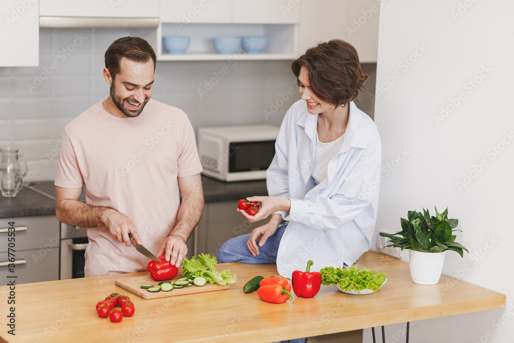 Funny young couple two friends guy girl in casual clothes sit on table preparing vegetable salad cooking food in light kitchen at home. Dieting family healthy lifestyle concept. Mock up copy space.