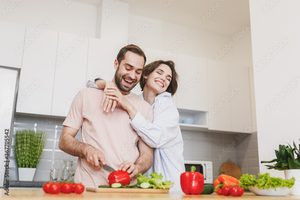 Smiling young couple two friends guy girl in casual clothes preparing vegetable salad cooking food in light kitchen at home. Dieting family healthy lifestyle concept. Mock up copy space. Hugging. Smiling young couple two friends guy girl in casual clothes preparing vegetable salad cooking food in light kitchen at home. Dieting family healthy lifestyle concept. Mock up copy space. Hugging.