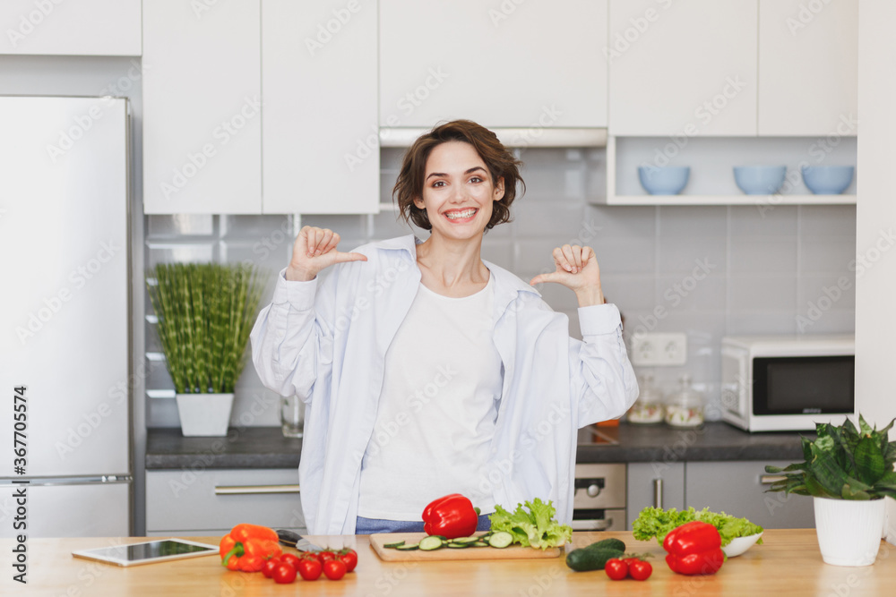 Smiling young housewife woman girl in casual clothes preparing vegetable salad cooking food in light kitchen at home. Dieting healthy lifestyle concept. Mock up copy space. Pointing thumbs on herself.