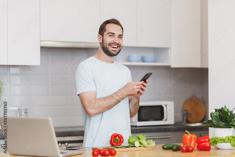 Funny young bearded man in white t-shirt using mobile phone looking for recipe preparing vegetable salad cooking food in light kitchen at home. Dieting healthy lifestyle concept. Mock up copy space. Funny young bearded man in white t-shirt using mobile phone looking for recipe preparing vegetable salad cooking food in light kitchen at home. Dieting healthy lifestyle concept. Mock up copy space.