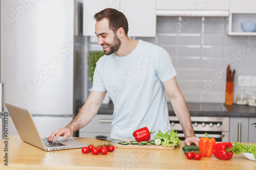 Smiling young man guy in casual t-shirt using laptop computer looking at recipe preparing vegetable salad cooking food in light kitchen at home. Dieting healthy lifestyle concept. Mock up copy space.