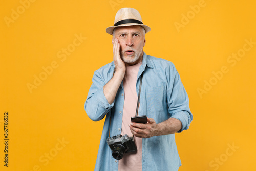 Amazed traveler tourist elderly gray-haired man isolated on yellow background. Passenger traveling abroad on weekends. Air flight journey. Using mobile cell phone booking taxi hotel put hand on cheek.