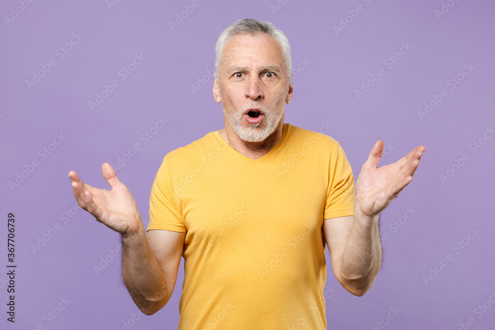 Shocked elderly gray-haired mustache bearded man in yellow t-shirt posing isolated on pastel violet background. People lifestyle concept. Mock up copy space. Keeping mouth open, spreading hands. Shocked elderly gray-haired mustache bearded man in yellow t-shirt posing isolated on pastel violet background. People lifestyle concept. Mock up copy space. Keeping mouth open, spreading hands.