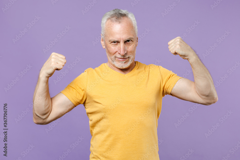 Strong elderly gray-haired mustache bearded man in casual yellow t-shirt posing isolated on pastel violet wall background studio. People lifestyle concept. Mock up copy space. Showing biceps, muscles. Strong elderly gray-haired mustache bearded man in casual yellow t-shirt posing isolated on pastel violet wall background studio. People lifestyle concept. Mock up copy space. Showing biceps, muscles.