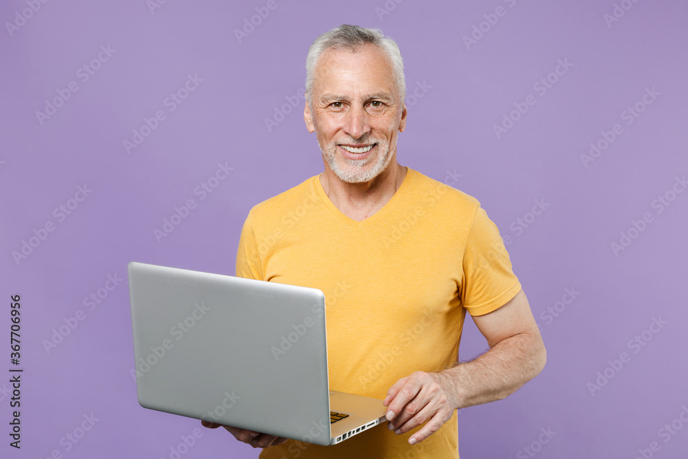 Smiling elderly gray-haired mustache bearded man in casual yellow t-shirt isolated on violet background studio portrait. People lifestyle concept. Mock up copy space. Working on laptop pc computer. Smiling elderly gray-haired mustache bearded man in casual yellow t-shirt isolated on violet background studio portrait. People lifestyle concept. Mock up copy space. Working on laptop pc computer.