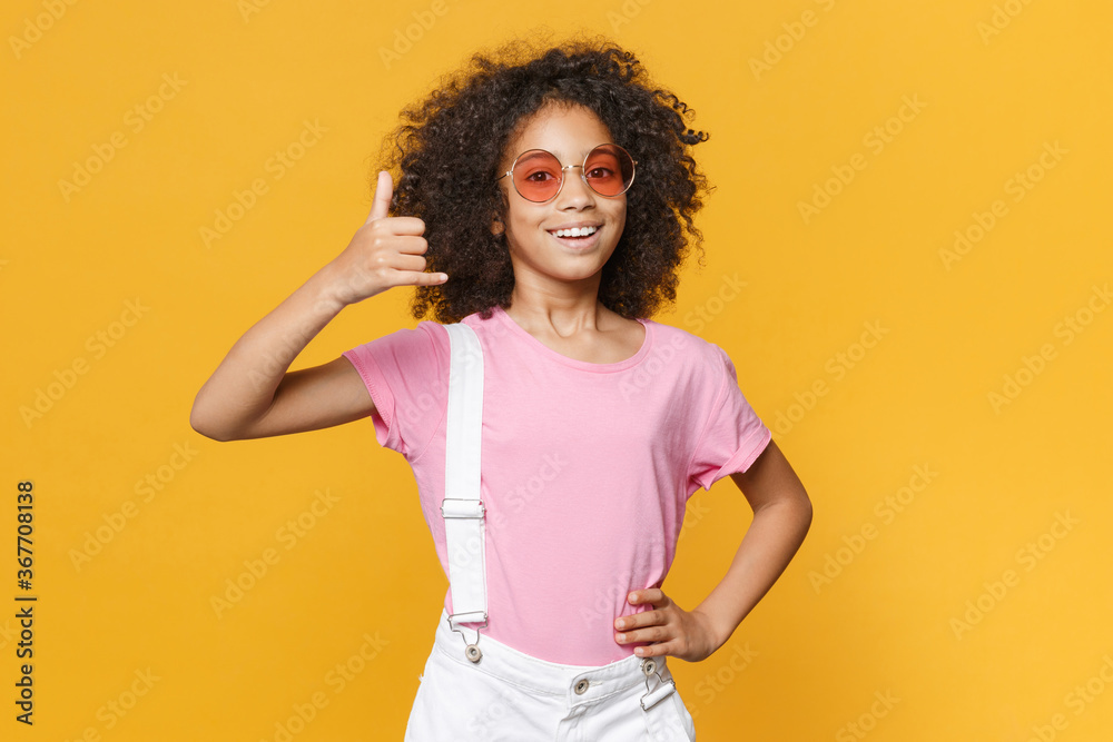 Smiling little african american kid girl 12-13 years old in pink t-shirt eyeglasses isolated on yellow background in studio. Childhood lifestyle concept. Doing phone gesture like says call me back. Smiling little african american kid girl 12-13 years old in pink t-shirt eyeglasses isolated on yellow background in studio. Childhood lifestyle concept. Doing phone gesture like says call me back.