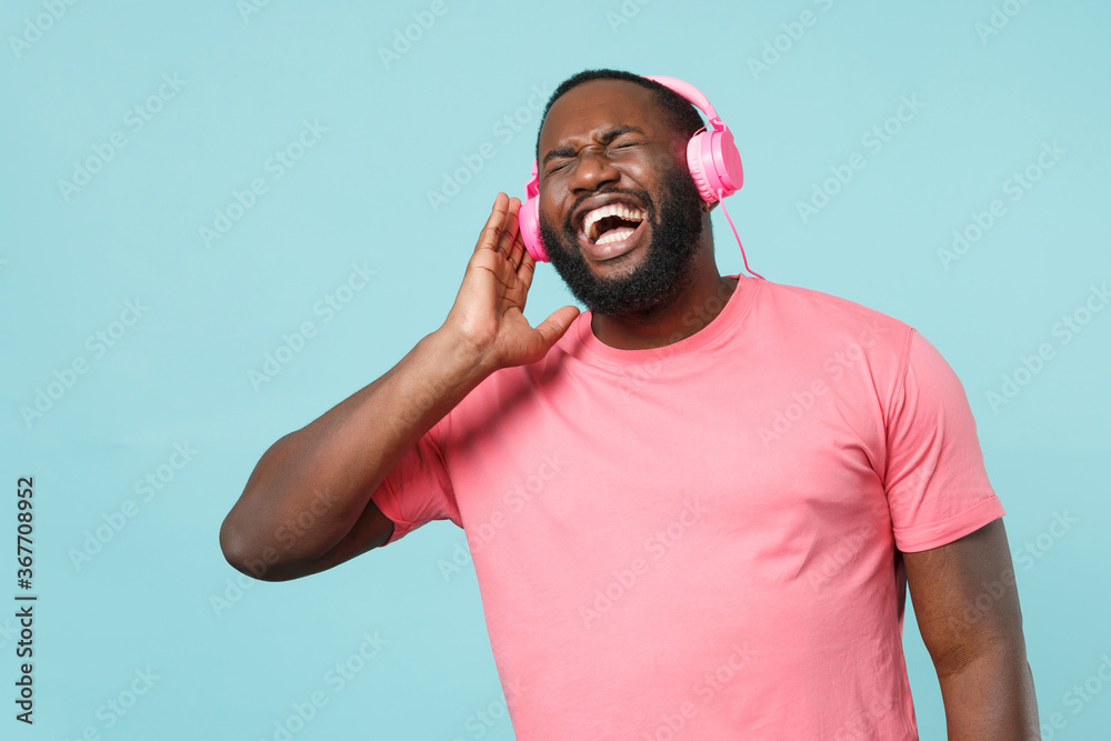 Naklejka premium Cheerful young african american man in casual pink t-shirt isolated on blue background studio portrait. People lifestyle concept. Mock up copy space. Listen music with headphones keeping eyes closed.