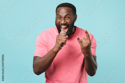 Funny young african american man guy in casual pink t-shirt isolated on blue background studio. People lifestyle concept. Mock up copy space. Depicting heavy metal rock sign, sing song in microphone.