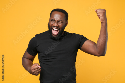 Overjoyed african american man guy football fan in casual black t-shirt isolated on yellow wall background studio portrait. People lifestyle concept. Mock up copy space. Clenching fists like winner.