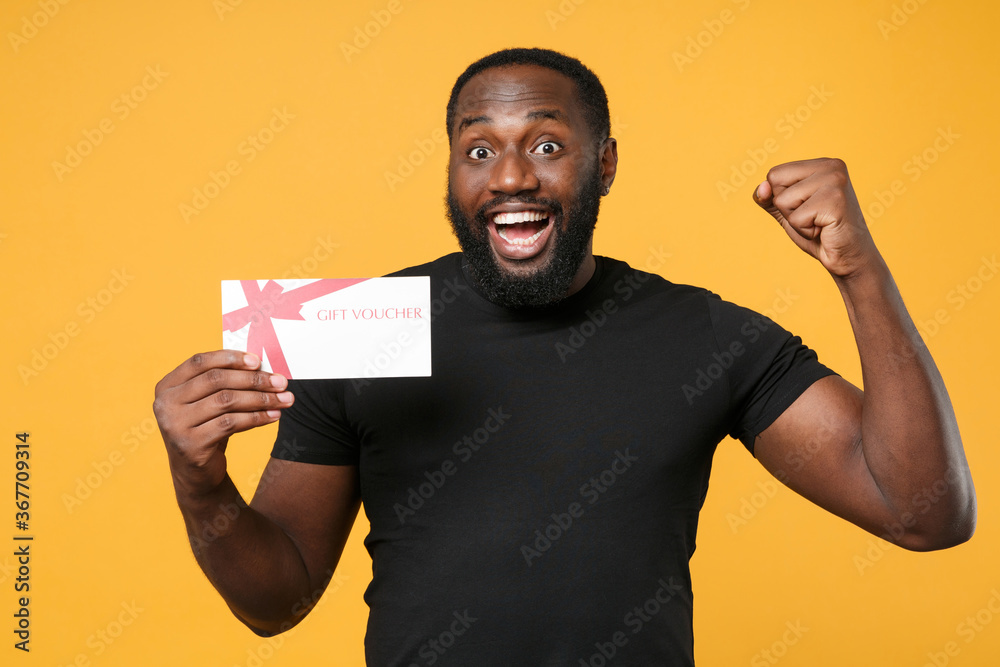 Excited african american man guy in casual black t-shirt isolated on yellow wall background studio portrait. People lifestyle concept. Mock up copy space. Hold gift certificate doing winner gesture. Excited african american man guy in casual black t-shirt isolated on yellow wall background studio portrait. People lifestyle concept. Mock up copy space. Hold gift certificate doing winner gesture.