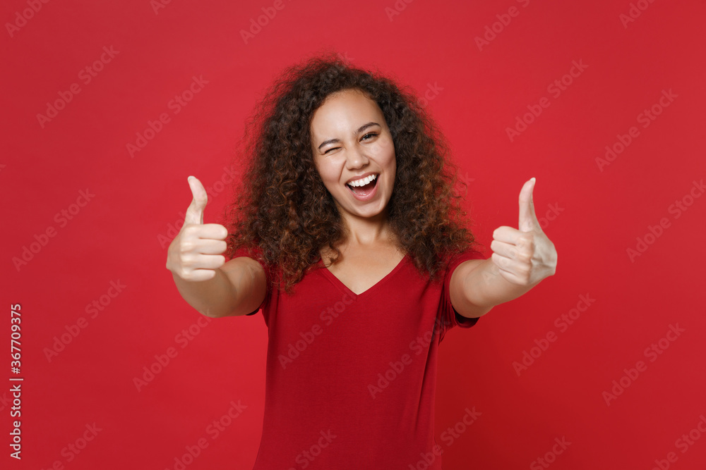 Blinking funny young african american woman girl in casual t-shirt posing isolated on red background studio portrait. People sincere emotions lifestyle concept. Mock up copy space. Showing thumbs up. Blinking funny young african american woman girl in casual t-shirt posing isolated on red background studio portrait. People sincere emotions lifestyle concept. Mock up copy space. Showing thumbs up.
