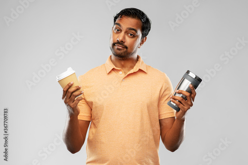 eco living and sustainability concept - young man in polo t-shirt comparing thermo cup or tumbler with disposable paper coffee cup over grey background