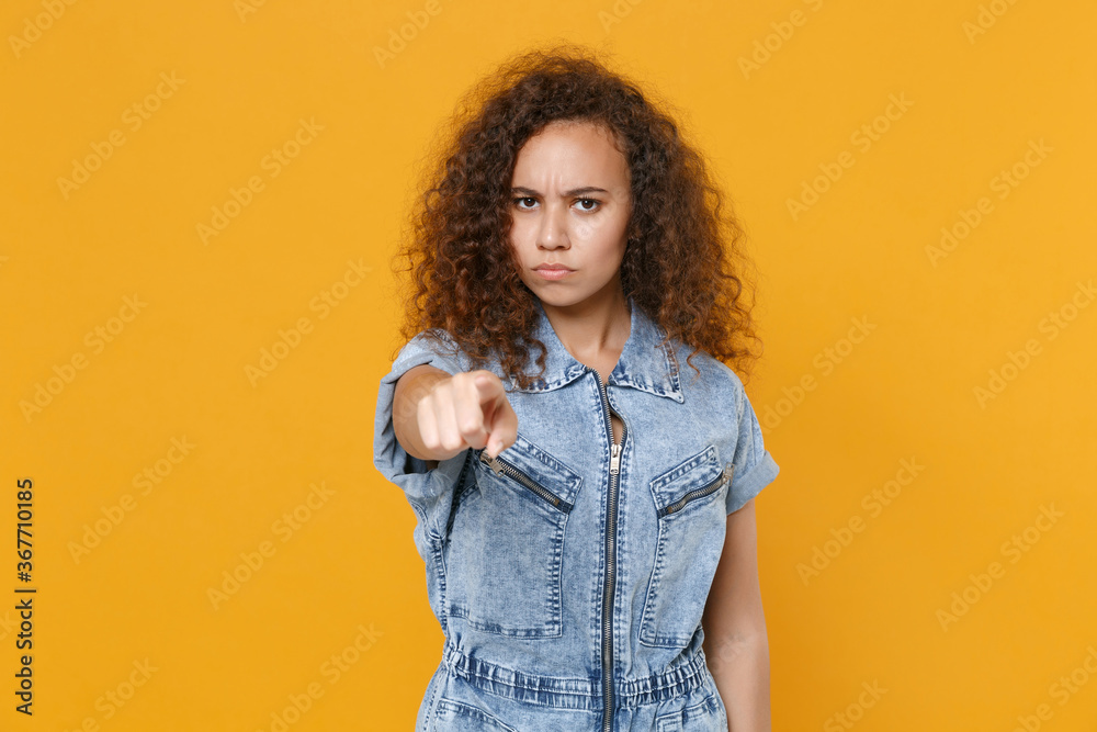 Displeased young african american woman girl in casual denim clothes isolated on yellow wall background studio portrait. People lifestyle concept. Mock up copy space. Pointing index finger on camera. Displeased young african american woman girl in casual denim clothes isolated on yellow wall background studio portrait. People lifestyle concept. Mock up copy space. Pointing index finger on camera.