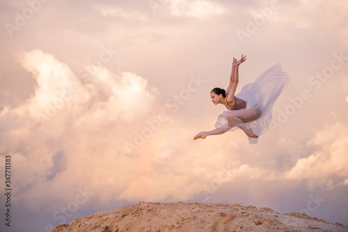 young ballerina in a light long dress soars above the ground, like a bird, in a dynamic jump against a background of pink sunset and clouds. A rainbow appears among the clouds.
