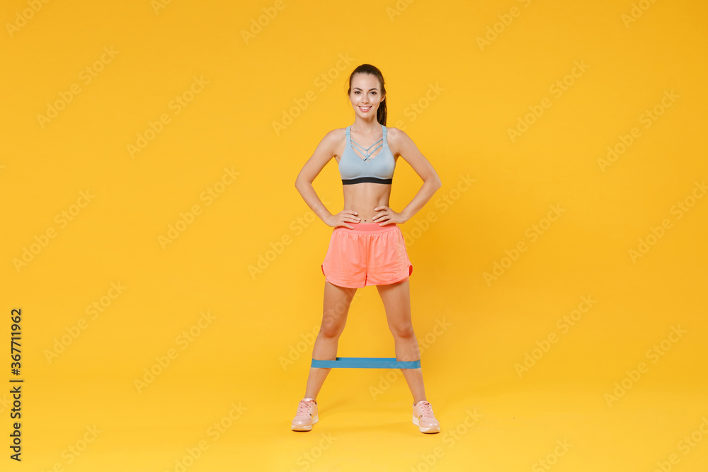 Full length portrait of smiling young fitness woman girl in sportswear working out isolated on yellow background. Workout sport motivation lifestyle concept. Doing exercise for legs with fitness gums. Full length portrait of smiling young fitness woman girl in sportswear working out isolated on yellow background. Workout sport motivation lifestyle concept. Doing exercise for legs with fitness gums.
