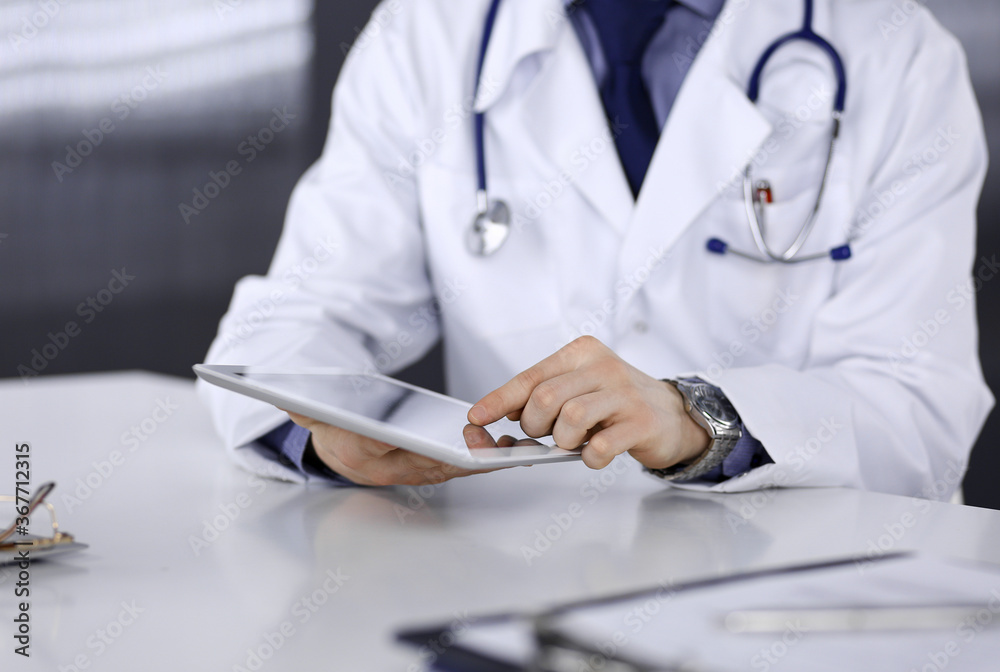 Fototapeta premium Unknown male doctor sitting and working with tablet computer in clinic at his working place, close-up. Young physician at work. Perfect medical service, medicine concept