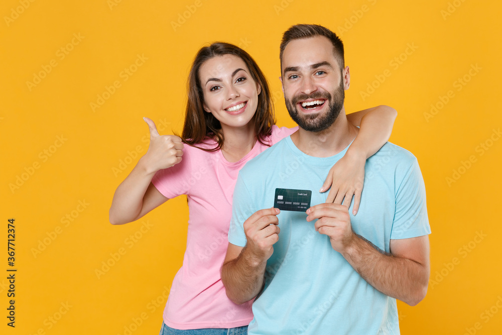 Funny young couple two friends guy girl in blue pink t-shirts posing isolated on yellow wall background. People lifestyle concept. Mock up copy space. Hold credit bank card, showing thumb up, hugging. Funny young couple two friends guy girl in blue pink t-shirts posing isolated on yellow wall background. People lifestyle concept. Mock up copy space. Hold credit bank card, showing thumb up, hugging.