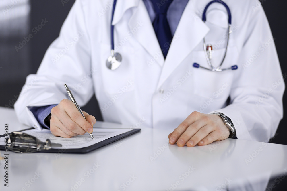 Unknown male doctor sitting and working with clipboard of medication history record in clinic at his working place, close-up. Young physician at work. Perfect medical service, medicine concept Unknown male doctor sitting and working with clipboard of medication history record in clinic at his working place, close-up. Young physician at work. Perfect medical service, medicine concept