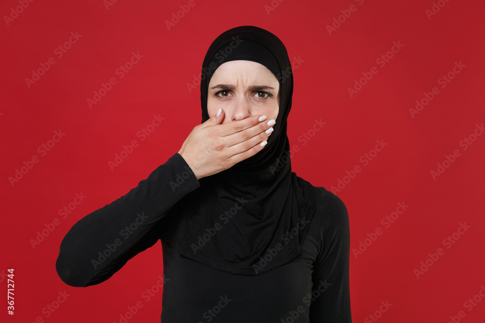 Displeased young arabian muslim woman in hijab black clothes posing isolated on bright red background studio portrait. People religious lifestyle concept. Mock up copy space. Covering mouth with hand. Displeased young arabian muslim woman in hijab black clothes posing isolated on bright red background studio portrait. People religious lifestyle concept. Mock up copy space. Covering mouth with hand.