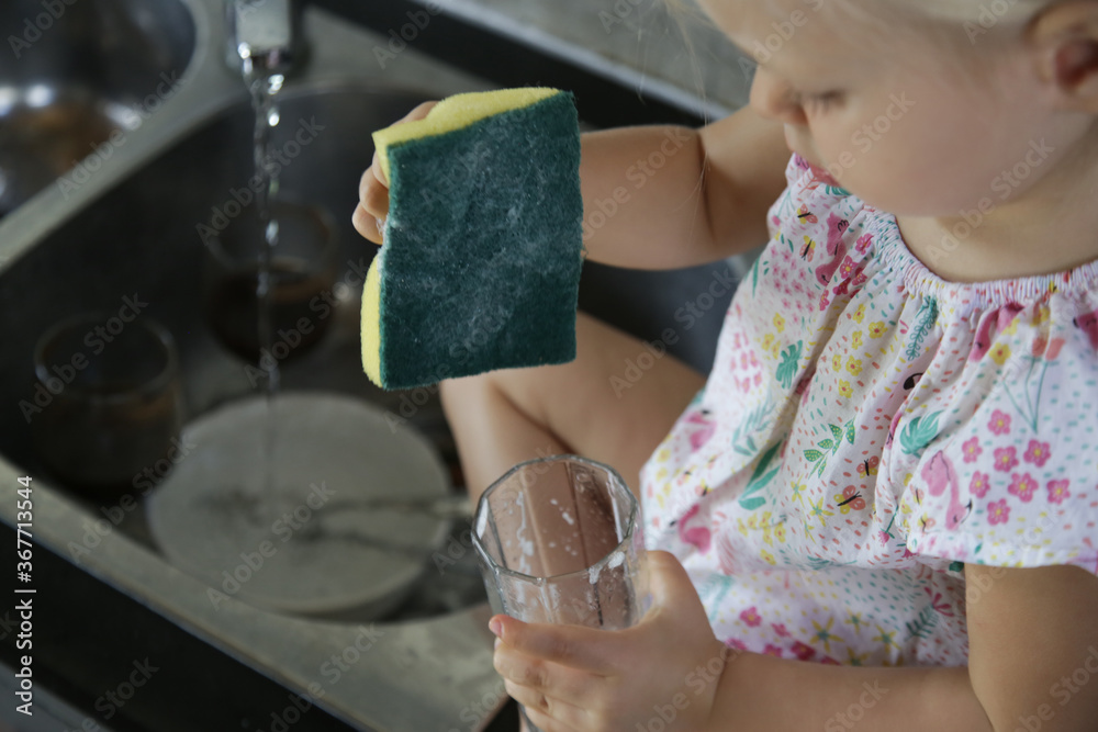 Child washing dishes in the kitchen. Cute two year old girl washes glass in sink. Early development, baby helps the parents. Chores for children. Child washing dishes in the kitchen. Cute two year old girl washes glass in sink. Early development, baby helps the parents. Chores for children.