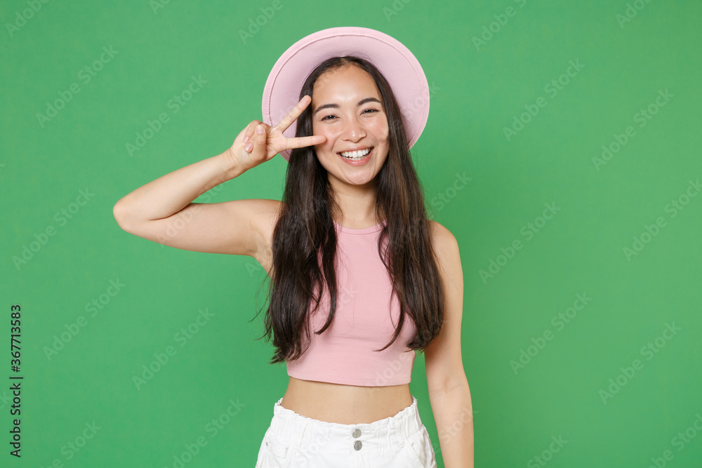 Smiling young asian woman girl in casual pink clothes hat isolated on green wall background studio portrait. People sincere emotions lifestyle concept. Mock up copy space. Showing victory sign. Smiling young asian woman girl in casual pink clothes hat isolated on green wall background studio portrait. People sincere emotions lifestyle concept. Mock up copy space. Showing victory sign.