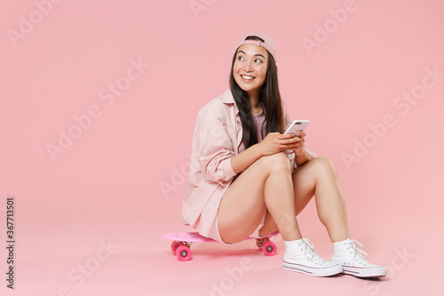 Full length portrait of smiling young asian woman girl in casual clothes, cap isolated on pastel pink background . People lifestyle concept. Sit on skateboard, using mobile cell phone, looking aside.