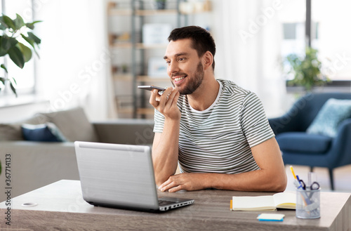 technology, remote job and people concept - happy smiling man with laptop computer calling on smartphone or using voice command recorder at home office