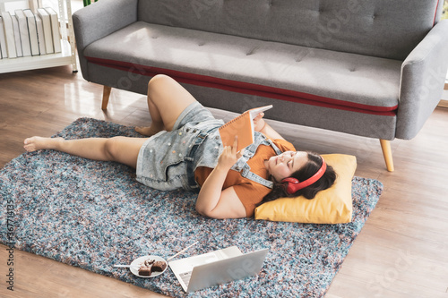 Fototapeta Plus size woman in headphone with computer notebook lying on carpet reading book