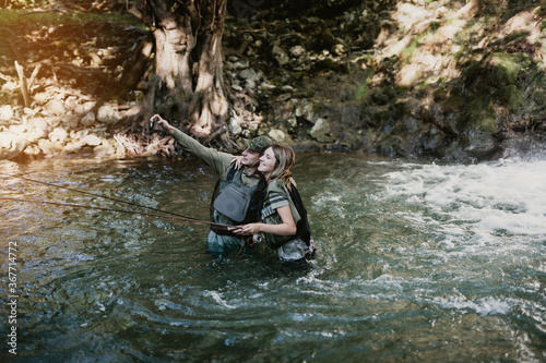 Fototapeta Naklejka Na Ścianę i Meble -  Young adult couple is fishing together on fast mountain river. They use smart phone to make selfie photo. Active people and sport fly fishing concept.