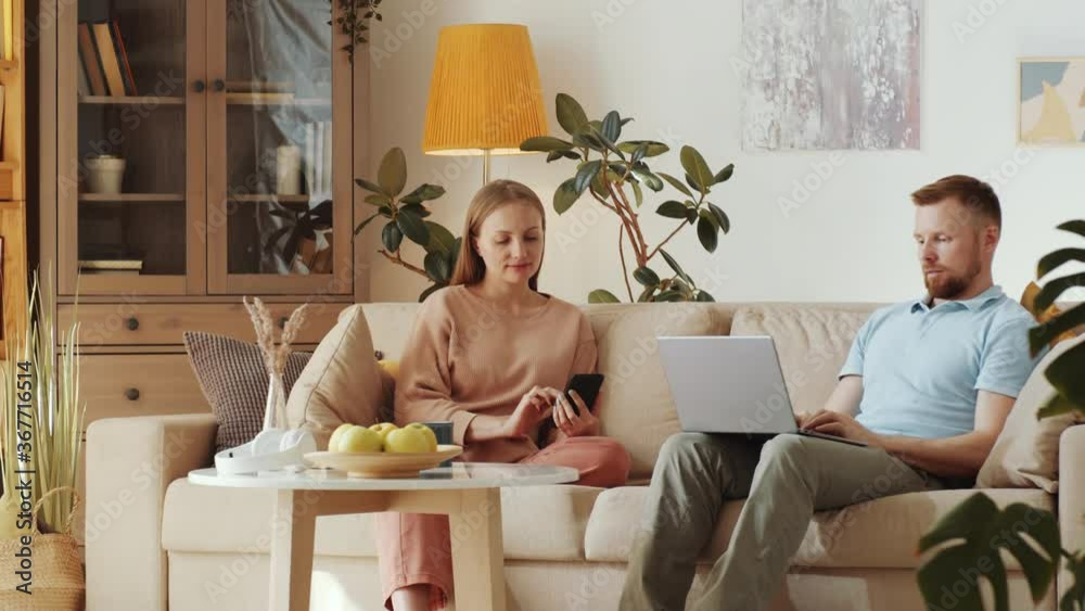 Young married couple sitting together on couch in the living room, woman typing on smartphone and man using laptop