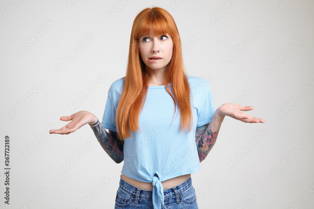 Embarrassed young redhead tattooed female biting her underlip while shrugging with raised palms, dressed in casual clothes while standing over white background Embarrassed young redhead tattooed female biting her underlip while shrugging with raised palms, dressed in casual clothes while standing over white background