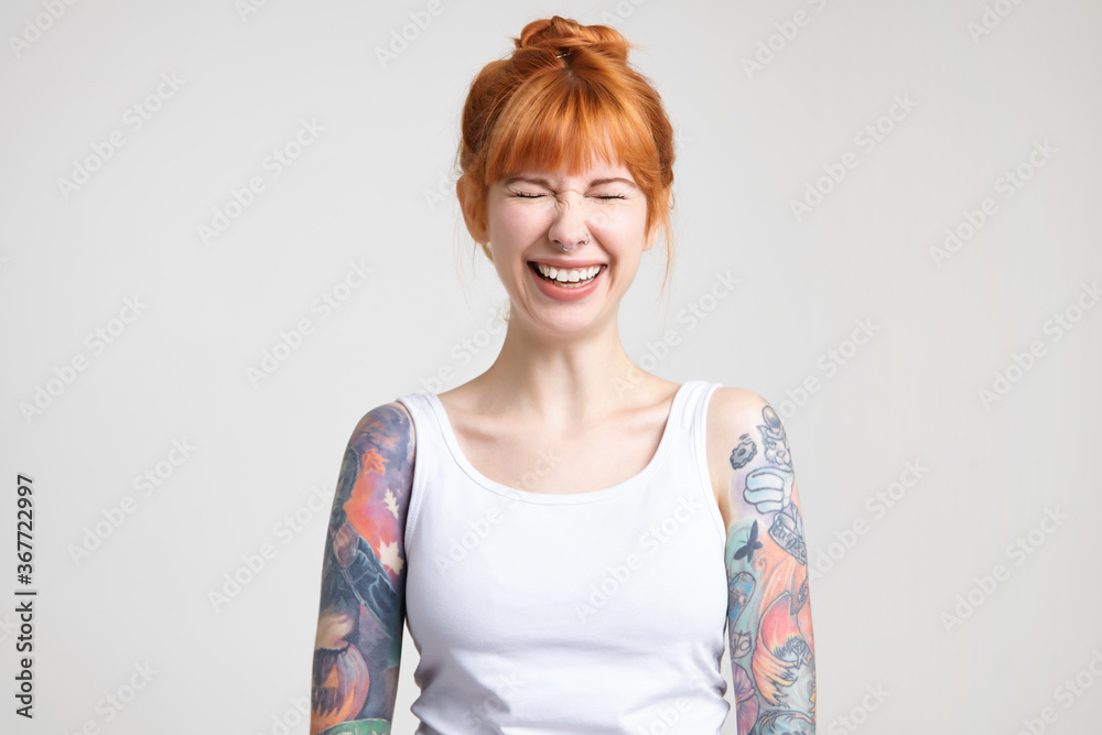 Joyful young attractive redhead woman with tattoos keeping her eyes closed while laughing cheerfully, dressed in white shirt while posing over white background Joyful young attractive redhead woman with tattoos keeping her eyes closed while laughing cheerfully, dressed in white shirt while posing over white background