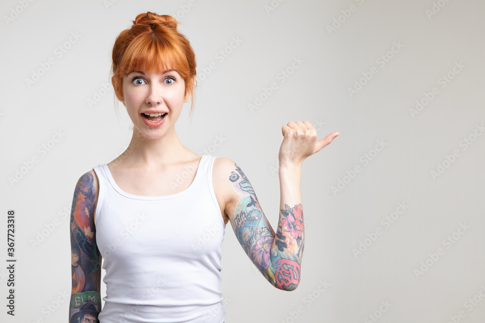 Excited young attractive redhead tattooed female rounding surprisedly her eyes while showing aside with raised hand, isolated over white background in casual shirt