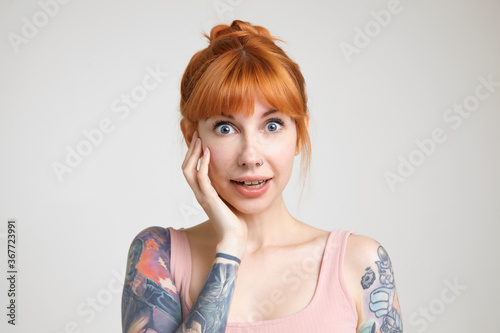 Portrait of young pretty surprised redhead lady with nose piercing keeping palm on her cheek while looking amazedly at camera, isolated over white background