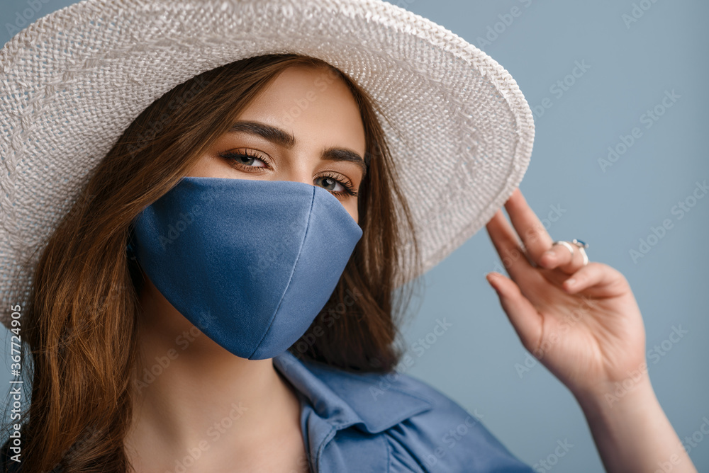 Woman wearing stylish protective face mask, summer white hat, posing on blue background. Trendy Fashion accessory during quarantine of coronavirus pandemic. Close up studio portrait.  - obrazy, fototapety, plakaty 