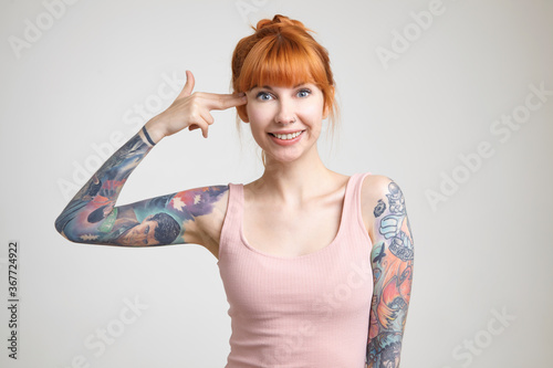 Indoor shot of young lovely redhead tattooed lady with bun hairstyle looking happily at camera and keeping hand with gun gesture near her temple  isolated over white background