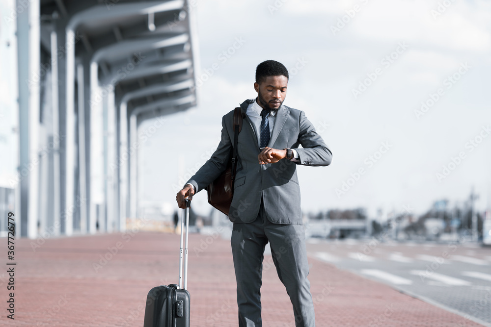 Φωτογραφία African Businessman With Travel Suitcase Looking At Watch Outside Airport