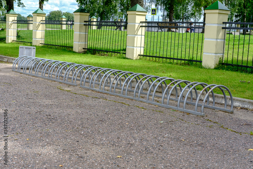 A bicycle parking in the park, made of painted metal arcs, rusty in places, stands on the asphalt against a backdrop of a metal fence and green grass. A bicycle parking in the park, made of painted metal arcs, rusty in places, stands on the asphalt against a backdrop of a metal fence and green grass.