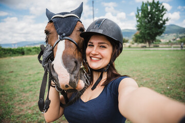 Beautiful young woman on a summer day taking a photo selfie with his horse in the grass field with the smartphone - Millennial has fun with his animal friend in a riding center