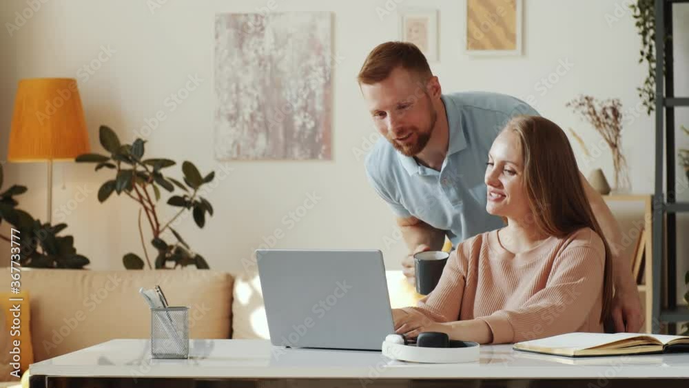 Beautiful woman using laptop at table in the living room while cheerful husband walking to her, drinking tea and discussing something on the Internet Beautiful woman using laptop at table in the living room while cheerful husband walking to her, drinking tea and discussing something on the Internet